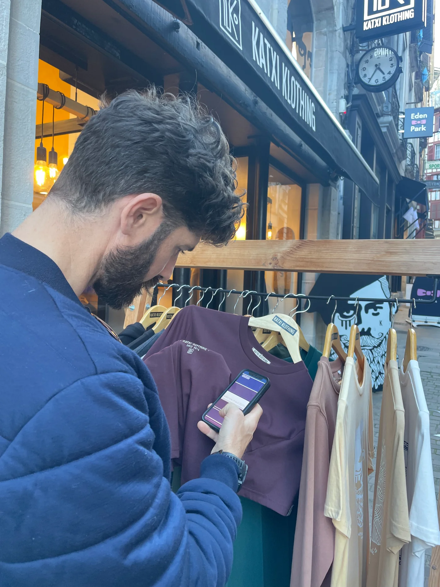 Un homme devant un portant de t-shirts consulte la palette digitale sur son téléphone pour choisir un vêtement en magasin