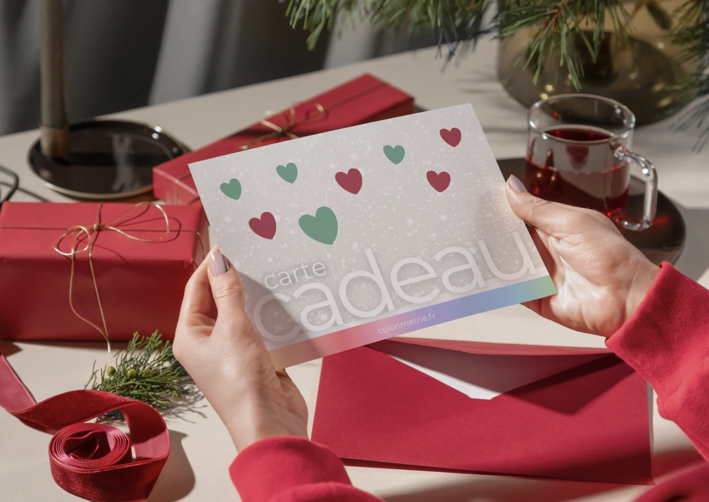 La carte cadeau Ihana édition Noël tenue entre deux mains, posée devant des cadeaux emballés de rouge, une tasse de thé et des branches de sapin